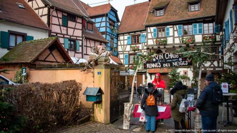Visitors beneath a “VIN CHAUD – RENNES DES NEIGES” sign at a Christmas stall surrounded by colorful half-timbered houses.