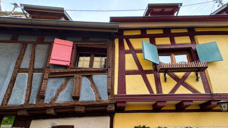 Blue-gray and yellow half-timbered façades with small windows, colorful shutters, and exposed diagonal braces.