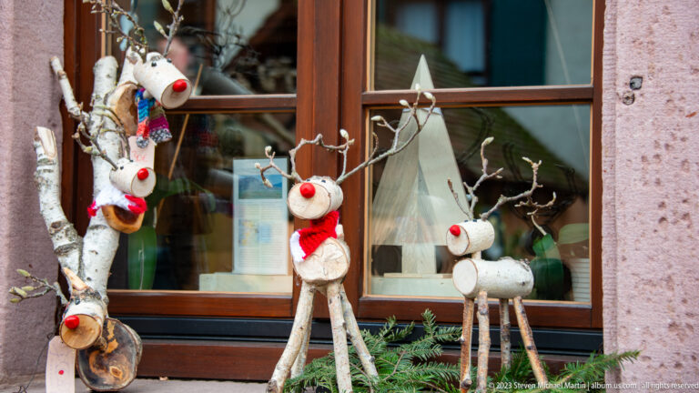 Three handmade birch-log reindeer with twig antlers, red noses, and scarves standing beside a wood-framed window.
