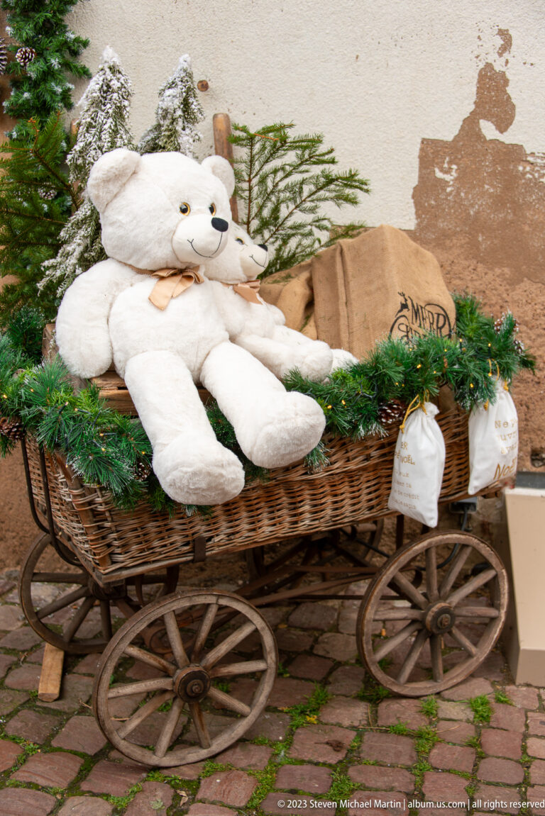 Large plush white bear reclining in a wooden spoked-wheel cart filled with evergreen Christmas greenery.