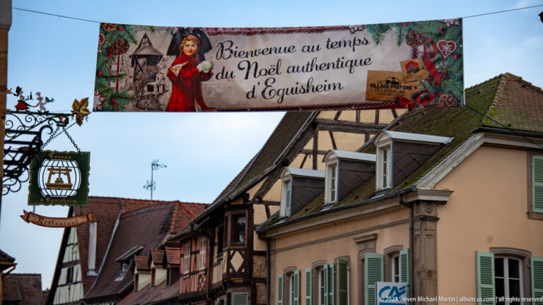 Banner reading “Bienvenue au temps du Noël authentique d’Eguisheim” hanging above historic village roofs.