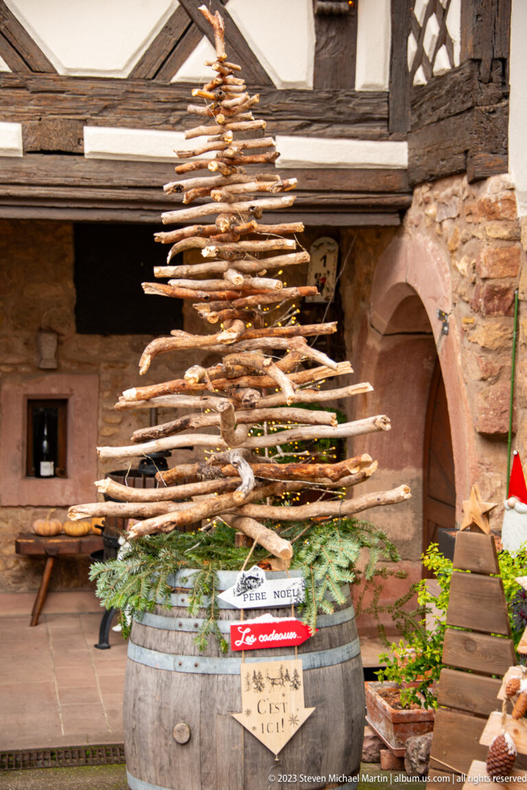 Christmas tree built from stacked branches on a wine barrel with signs for Père Noël, gifts, and “C’est ici.”