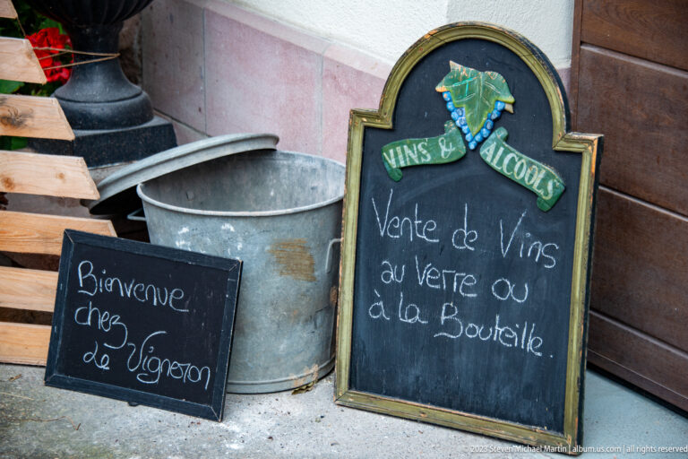 Chalkboards beside a metal tub welcoming visitors to a winegrower and advertising wine by glass or bottle.