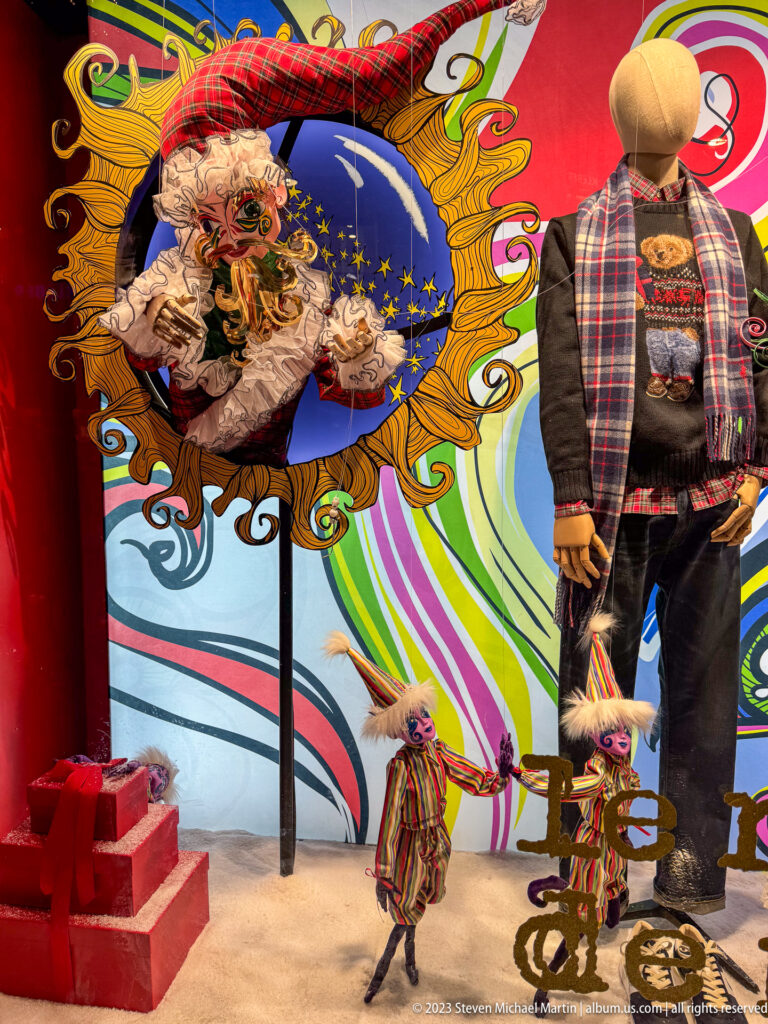Colorful holiday window with dressed mannequin, masked ruffled figure, and small puppet-like characters.