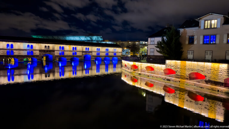 Barrage Vauban with blue-lit arches opposite a curved wall with red illuminated openings reflected in the river.