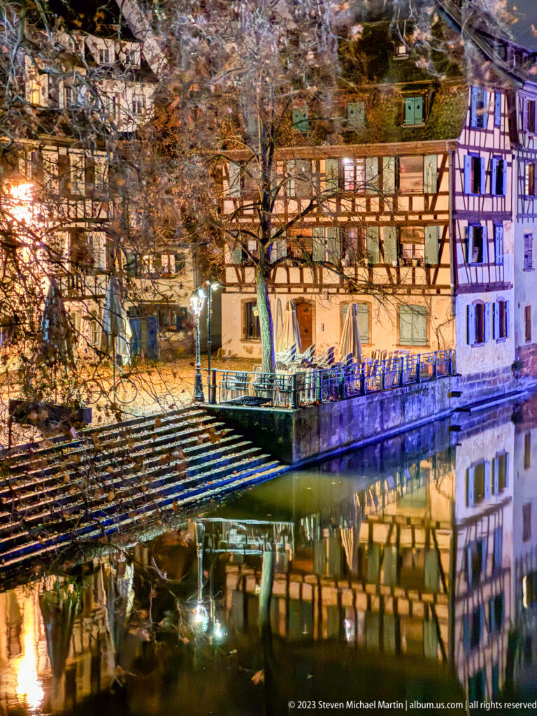 Half-timbered Petite France houses beside a canal, with stepped embankment, bare trees, and reflections at night.