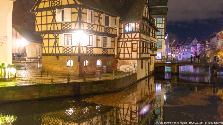Large half-timbered house beside a canal in Petite France, with more historic buildings and a small bridge beyond.