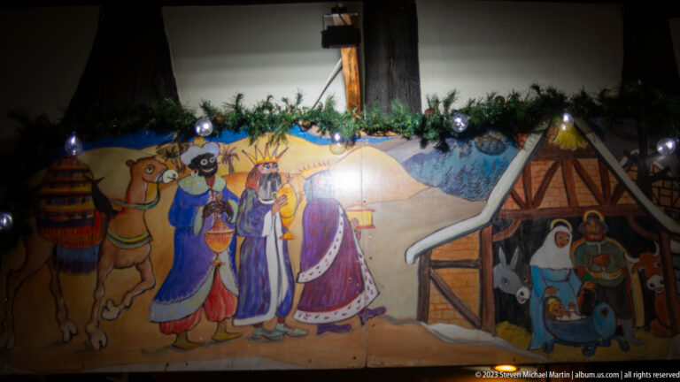 Painted Nativity scene with three Magi, camel, stable, Mary, Joseph, and infant Jesus beneath evergreen garland.