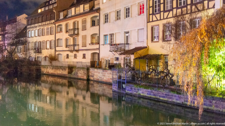 Pale canal-side houses with shuttered windows, balconies, and a lit weeping tree reflected in greenish water.