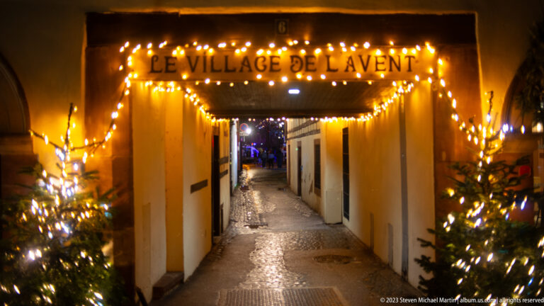 Narrow lit passage marked “Le Village de l’Avent,” framed by small Christmas trees and warm string lights.