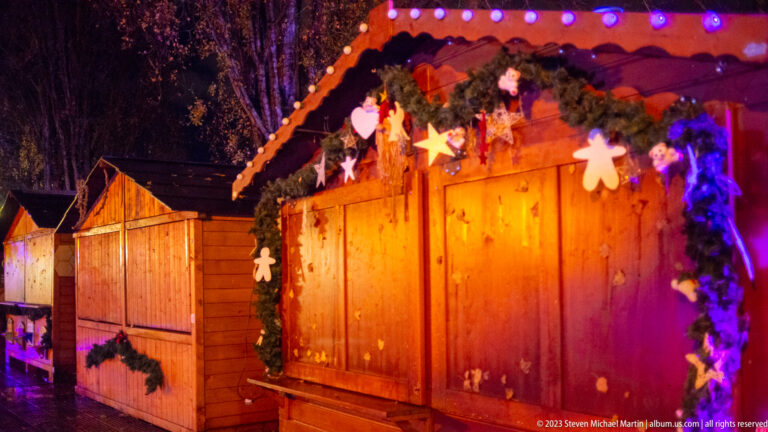 Row of closed wooden Christmas-market huts decorated with garlands, gingerbread figures, stars, and colored lights.