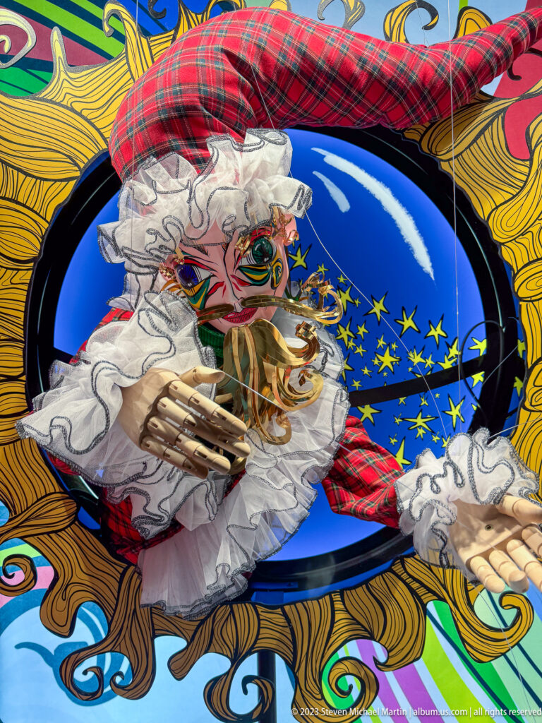 Close-up of a masked theatrical figure with white ruffles, tartan accents, painted face, and extended hand.