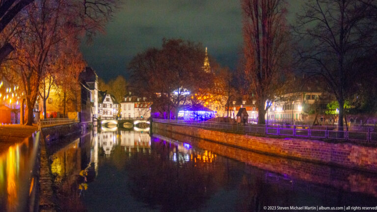 Strasbourg canal at night with historic buildings, low bridges, trees, and multicolored Christmas-light reflections.