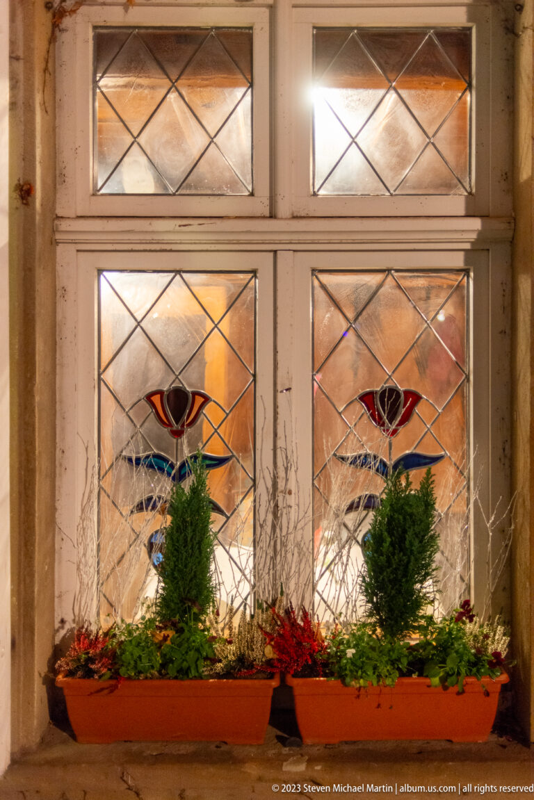 Two leaded windows with stylized colored-glass eye motifs above a planter holding small evergreens and red decorations.