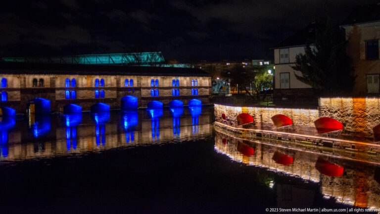 Barrage Vauban with blue-lit arches and neighboring red-lit embankment reflected in dark water.