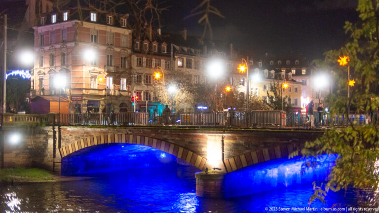 Low stone bridge glowing blue beneath its arch, with historic buildings and Christmas star lights beyond.