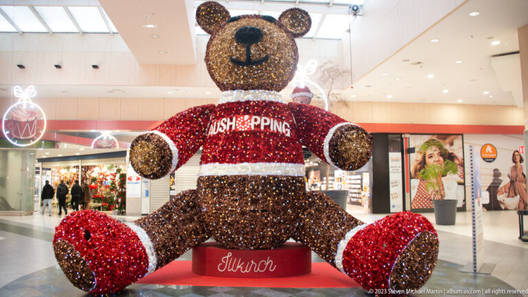 Giant sparkling teddy bear wearing a red “Aushopping” sweater in a bright shopping-center atrium.