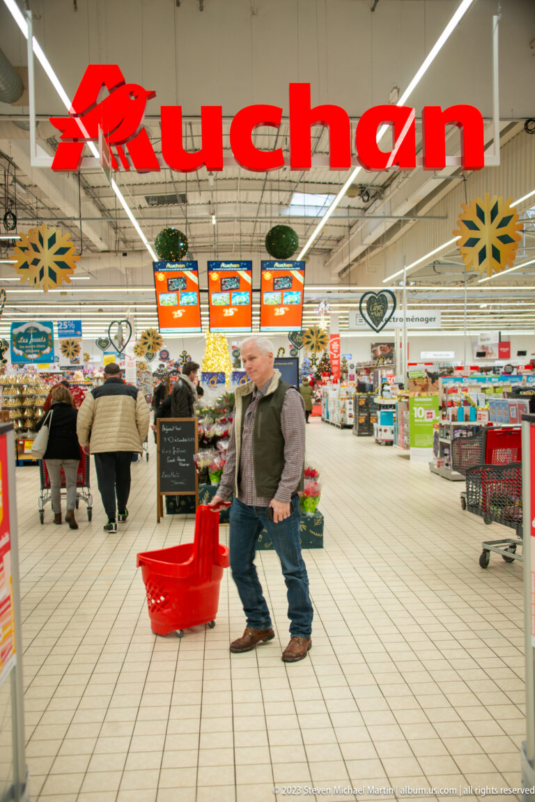 Auchan hypermarket interior with shoppers, large red store sign, promotional boards, and stacked red baskets.