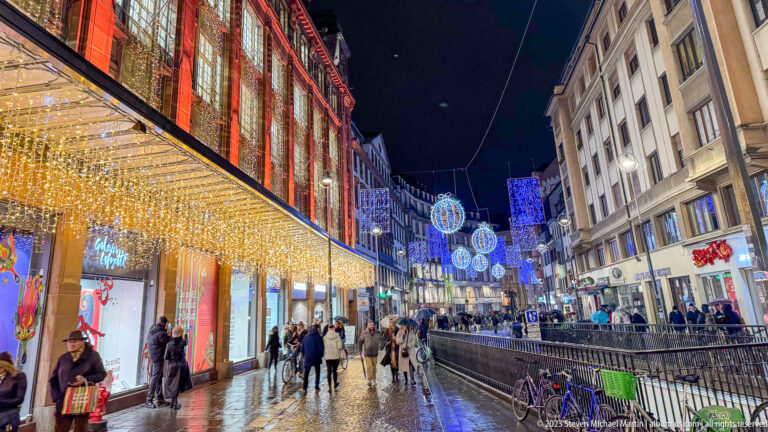 Wet Strasbourg shopping street with pedestrians beneath blue-and-white hanging Christmas ornaments and light curtains.