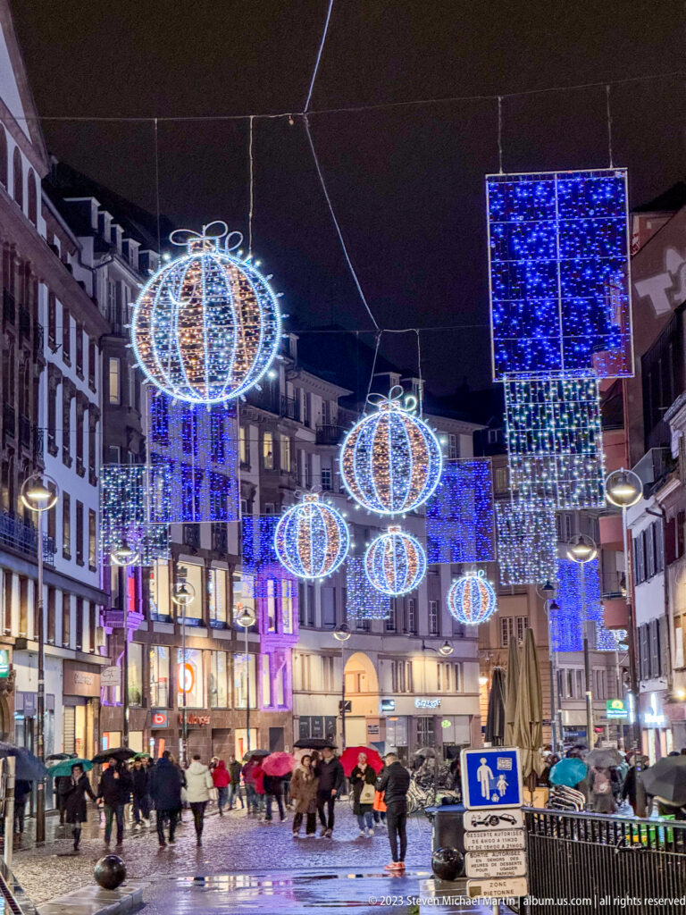 Wide Strasbourg intersection beneath glowing blue-and-white baubles and rectangular Christmas light panels.