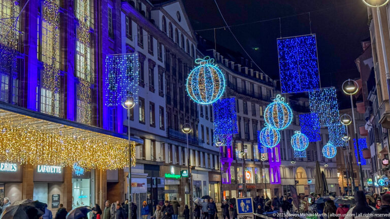 Galeries Lafayette frontage beside a crowded street hung with blue-and-white Christmas baubles and light panels.