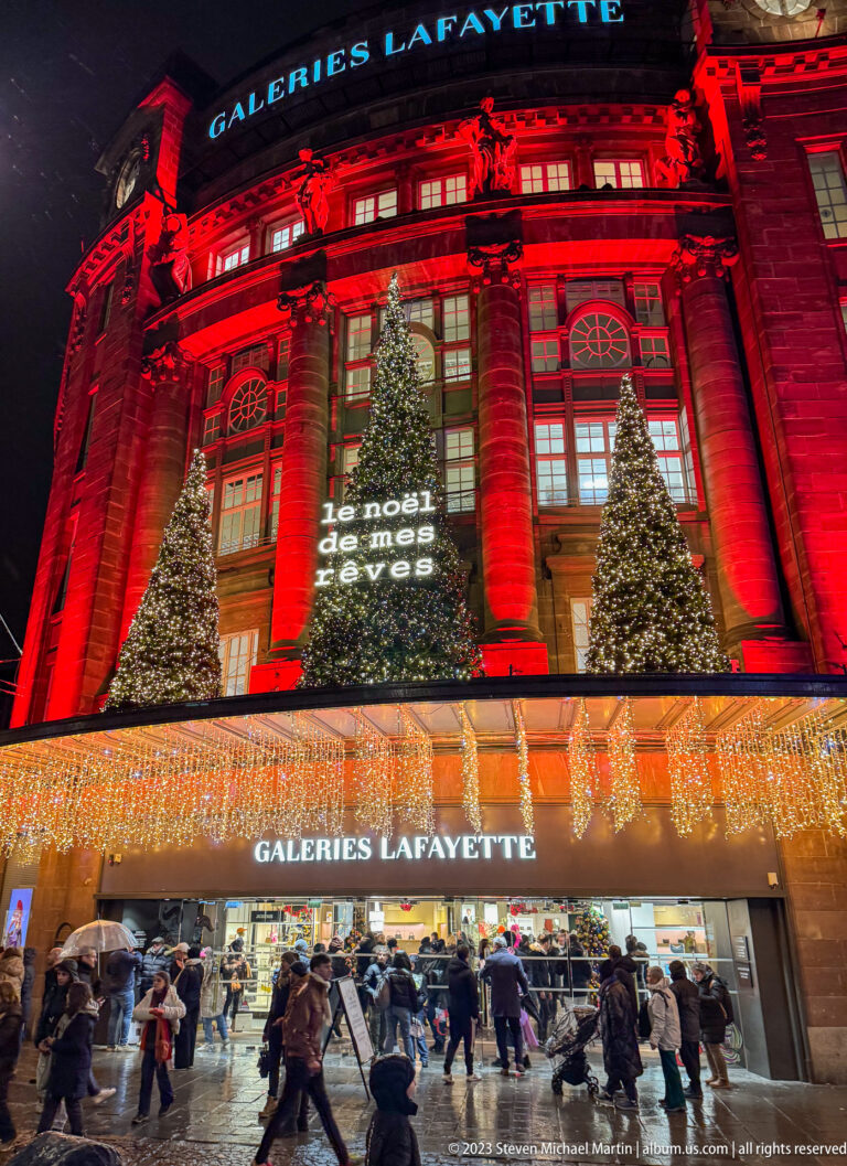 Galeries Lafayette Strasbourg glowing red with Christmas trees, warm storefront lights, and pedestrians on wet pavement.