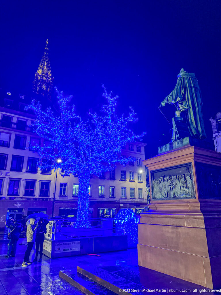 Statue of General Kléber on a stone pedestal beside a tree glowing blue in Place Kléber at night.