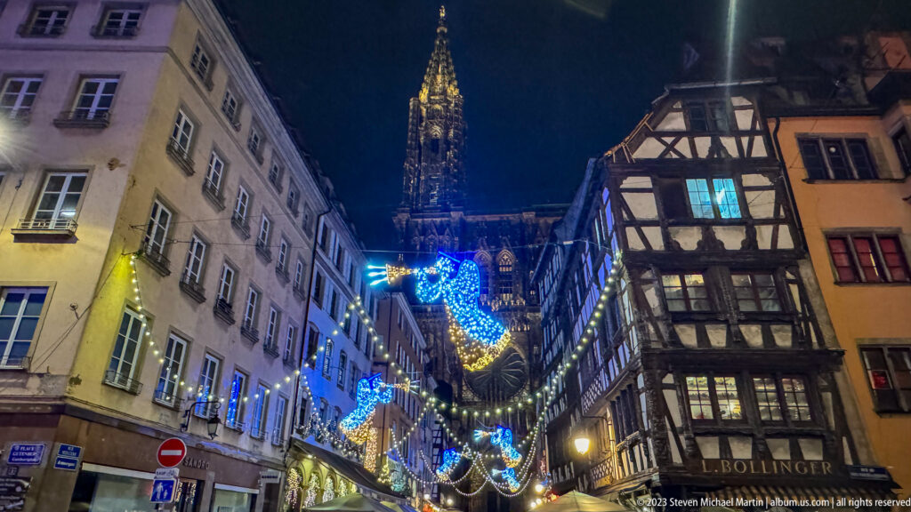 Strasbourg Cathedral spire above a narrow historic street with half-timbered building and blue-and-gold Christmas lights.