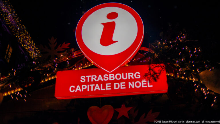 Red illuminated sign reading “Strasbourg Capitale de Noël” among dark greenery and small Christmas lights.