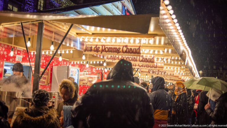 Rainy crowd at a bright market food stall advertising hot apple juice, with steam rising behind the counter.
