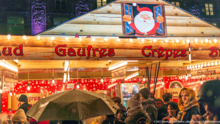 Christmas-market chalet marked “Gaufres” and “Crêpes,” with Santa decoration and customers holding umbrellas.