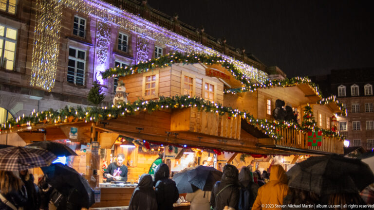 Large two-story wooden Christmas chalet with garlands, balcony, lights, serving counter, and visitors under umbrellas.