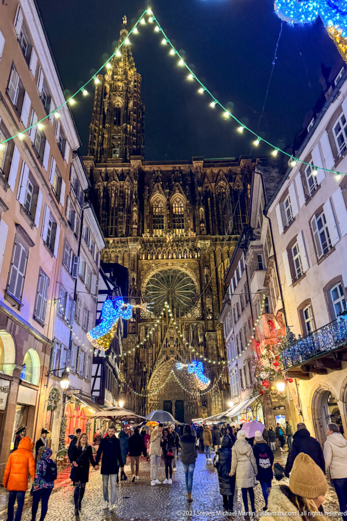 Strasbourg Cathedral west façade and rose window above a crowded wet street with blue Christmas lights.