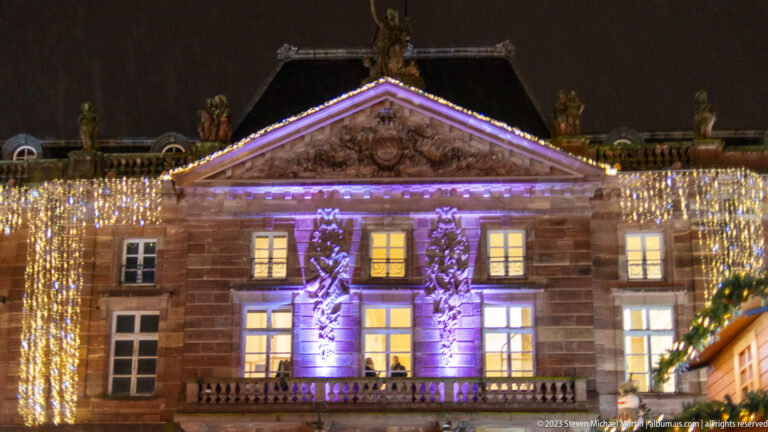Symmetrical sandstone building with classical pediment, purple uplighting, and dense curtains of warm Christmas lights.