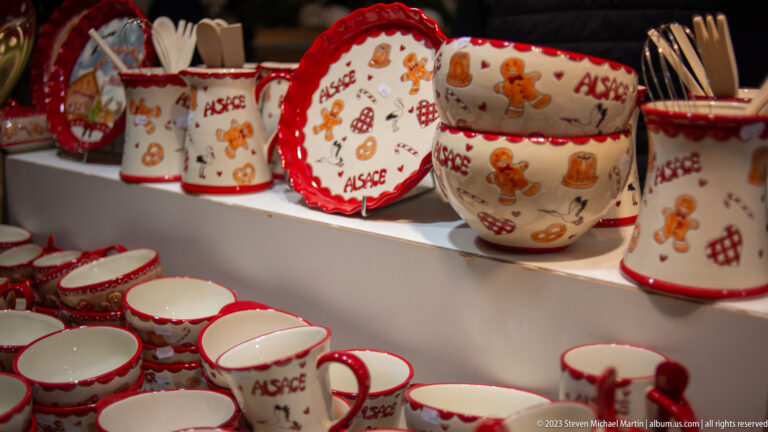 Red-and-white mugs, bowls, and plates decorated with gingerbread figures and the word “Alsace.”