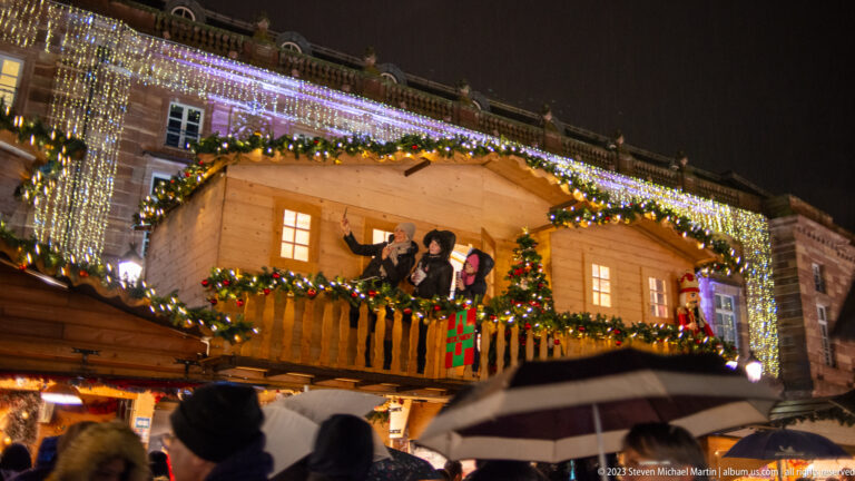 Decorated two-story Christmas chalet with costumed figures on the balcony and umbrellas in the foreground.