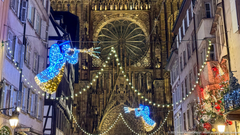 Strasbourg Cathedral rose window framed by narrow street façades, string lights, and large blue angel decorations.