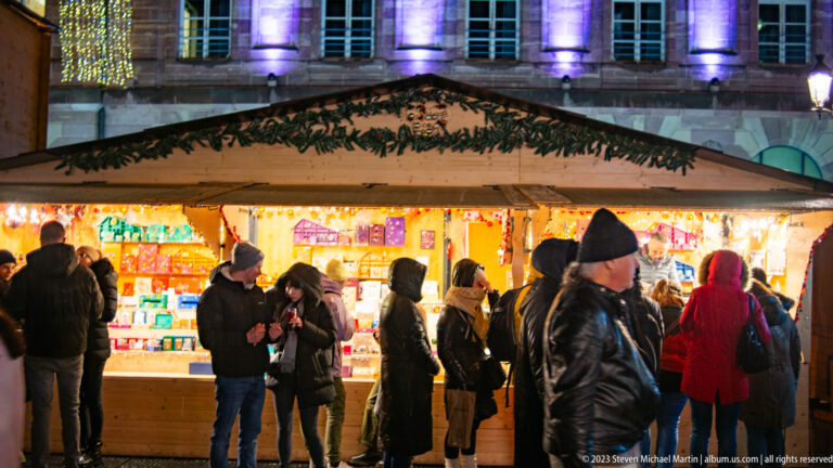 Shoppers browsing a brightly lit wooden Christmas-market stall filled with colorful merchandise.