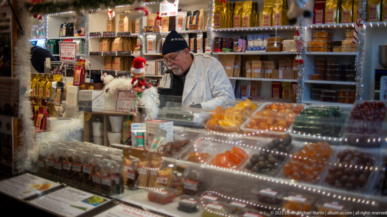 Vendor behind a glass case of sweets and confectionery at a crowded Christmas-market food stall.