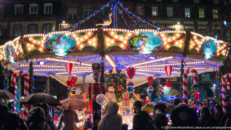 Crowd with umbrellas around a brightly lit Christmas carousel decorated with candy-cane columns and painted winter scenes.