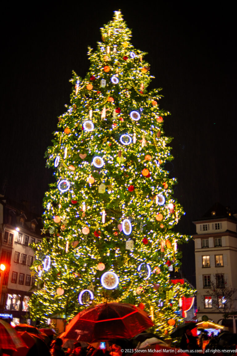 Large illuminated Place Kléber Christmas tree above red umbrellas on a rainy night.