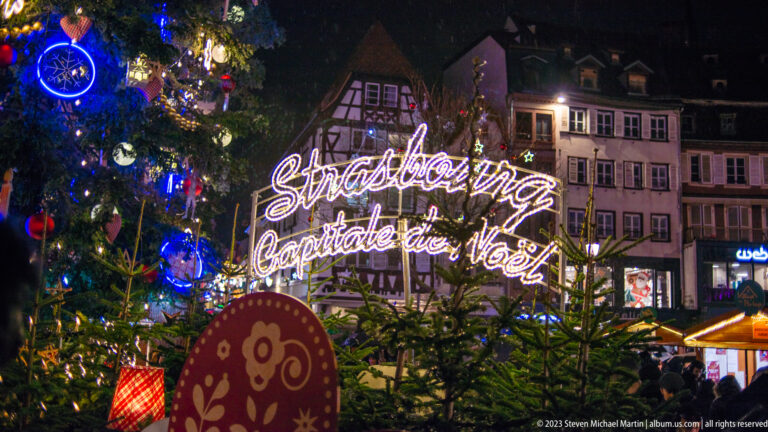 Glowing “Strasbourg Capitale de Noël” script above evergreen trees with historic façades behind.
