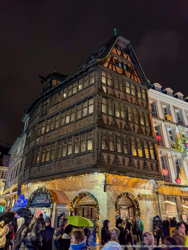 Illuminated Maison Kammerzell with carved timber framing, leaded windows, steep gable, and market crowd below.
