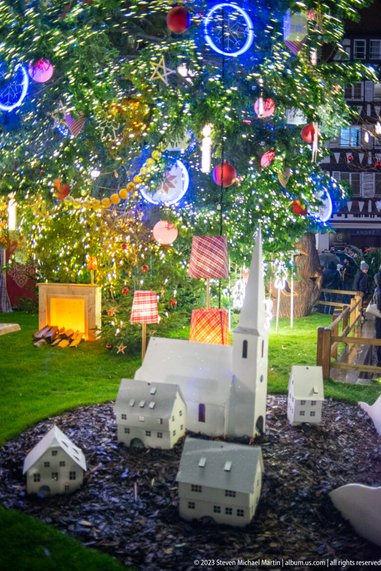 Miniature white church and houses arranged beneath a Christmas tree with glowing ornaments and gift decorations.