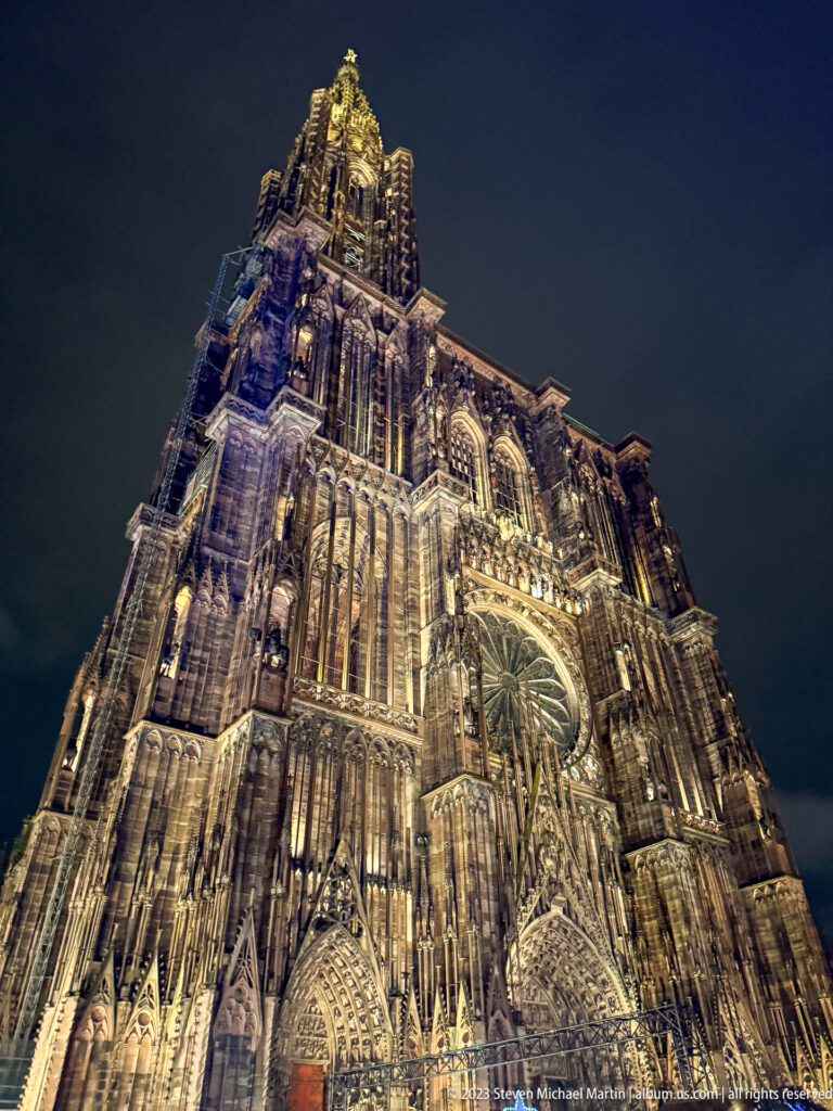 Low-angle night view of Strasbourg Cathedral’s west façade with rose window, portals, and dense Gothic tracery.