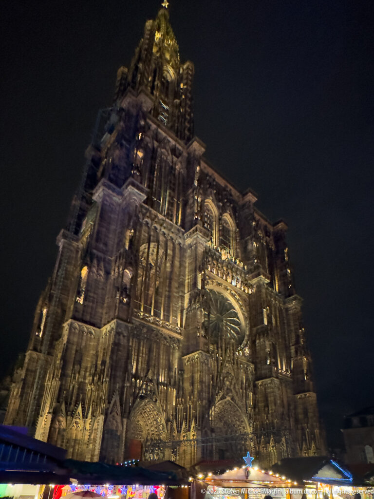 Strasbourg Cathedral’s illuminated west façade rising above Christmas-market chalet roofs and festive lights.