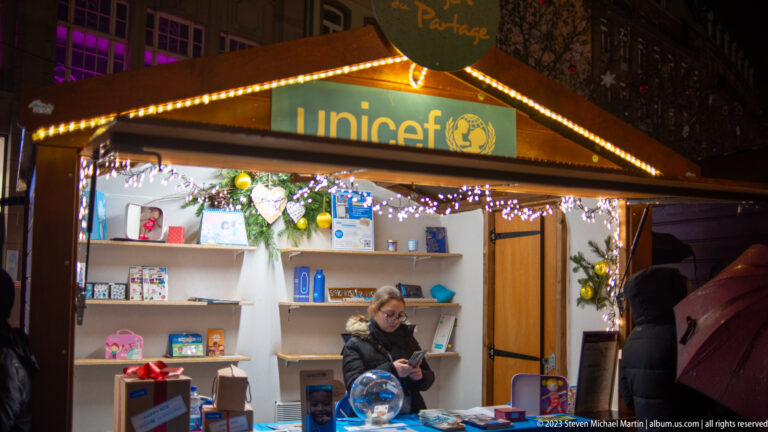 UNICEF Christmas-market stall with gifts, evergreen decoration, white lights, and a vendor behind the counter.