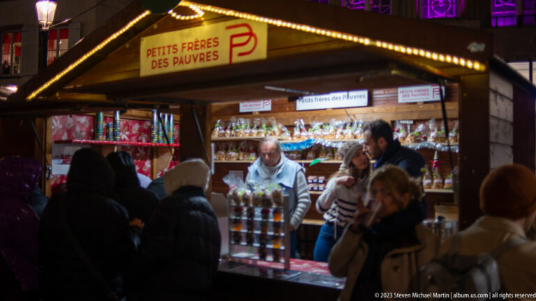 Petits Frères des Pauvres market chalet with vendor, jars, packaged sweets, and customers gathered in front.