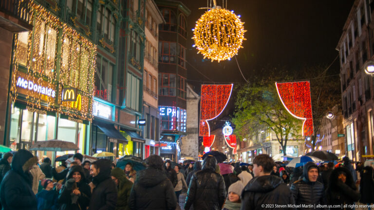 Crowded Strasbourg shopping street with umbrellas beneath a glowing gold sphere and red Christmas light panels.