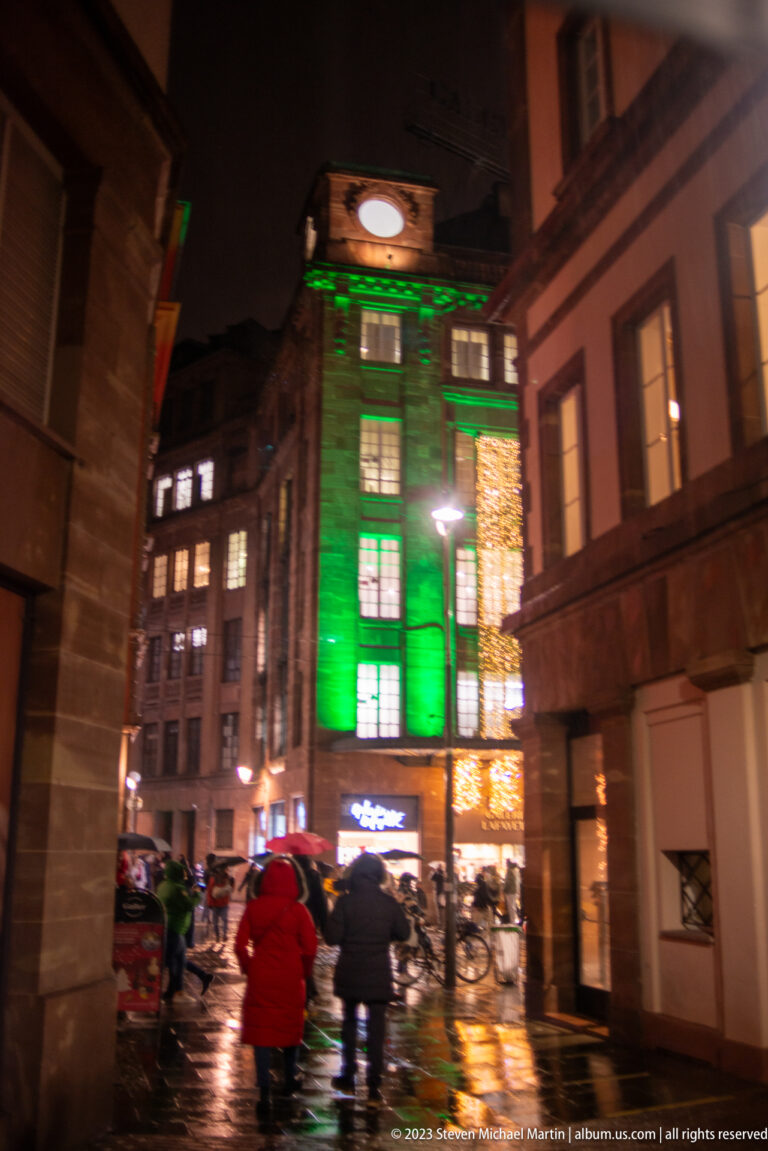 Historic Strasbourg building illuminated green beyond wet pavement, umbrellas, and neighboring warm Christmas lights.